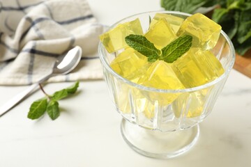 Yellow jelly cubes in dessert bowl and mint on white marble table, closeup
