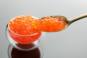 Taking red caviar with spoon from bowl at grey mirror surface, closeup