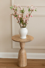 pink cherry blossom branches in a white ceramic vase sitting on a wooden side table against a cream colored wall with wainscotting, room for copy