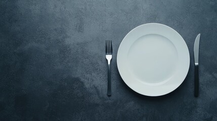 Centered on a gray stone surface, a white ceramic plate waits for culinary delights, flanked by a fork and knife in a refined minimalist layout