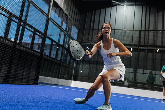 Focused woman playing padel tennis on indoor court, preparing to hit ball, copy space