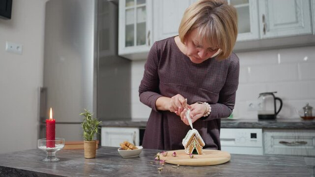 Woman bent over, carefully decorating freshly baked cookies with intricate icing details, wooden tray holds gingerbread house, surrounded by dried flowers, candle, and bowl of cookies