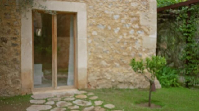 Blurry view of rustic mediterranean house exterior with stone wall and wooden door among lush greenery and small pathway creating peaceful atmosphere