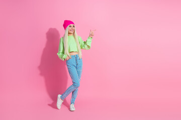 Stylish blonde woman in vibrant attire and hot pink beanie against a pink background pointing with energy and joy