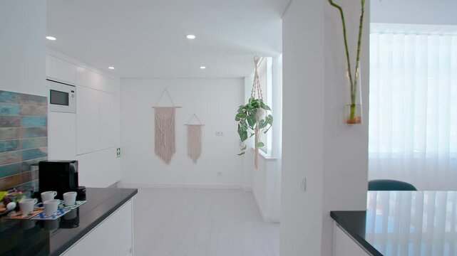 A modern kitchen with a glossy black countertop, a blue-tiled backsplash, open shelves with decor, hanging pendant lights, and macrame wall art in a bright interior space.