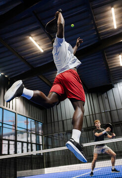 African American man jumping to hit ball during intense padel tennis match indoors