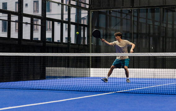 Young man playing padel tennis on blue court, focusing on hitting ball, copy space