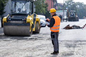 Construction worker conducts site inspection while monitoring roadwork with heavy machinery in...