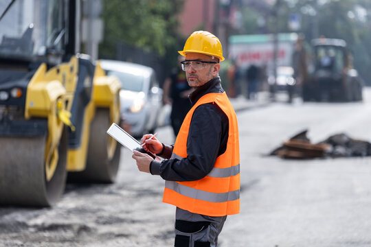 Construction worker supervises road work while taking notes during a busy construction project in an urban area