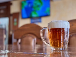 A Refreshing Glass of Beer is Poured on a Wooden Table, Perfectly Set for Enjoyment and Socializing. A frothy mug of icecold beer sits atop a beautifully polished wooden table, ideal for relaxation