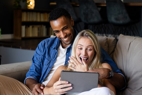 Celebrating engagement, diverse couple on couch with tablet, smiling and surprised