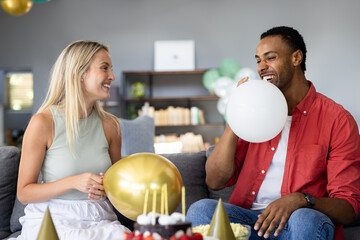 Smiling diverse couple inflating balloons at home for festive party, feeling joyful