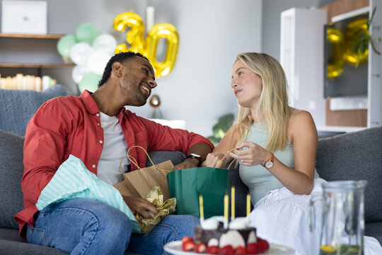 At party, Diverse couple celebrating birthday on couch with gifts and cake, smiling joyfully, at hom
