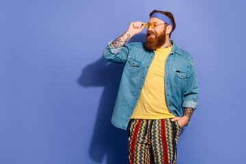 Fototapeta premium Trendy man with red beard wearing casual denim shirt and retro pants against a violet background, expressing happiness and confidence.
