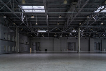 Industrial interior expanse, expansive space under a metal beam framework roof illuminated by...