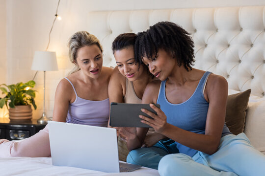 Diverse female friends relaxing on bed, using tablet and laptop, enjoying leisure time together