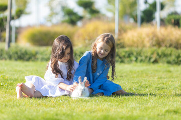 Fototapeta premium Happy little girls sitting on green grass and playing with white rabbit in summer day in park outdoor. High quality photo. Children and animals friendship and love concept. Copy space