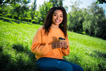 Photo of shiny cute girl dressed orange sweatshirt sitting grass enjoying beverage outdoors urban city park