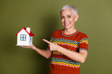 Smiling mature woman holding miniature house model on green background promoting real estate or...