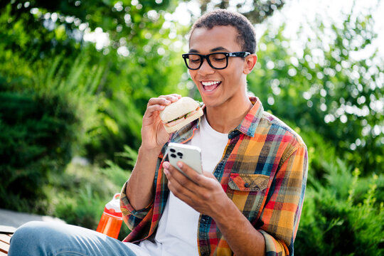 Photo of nice young man use smart phone eat sandwich wear checkered shirt city center park fresh air outdoors