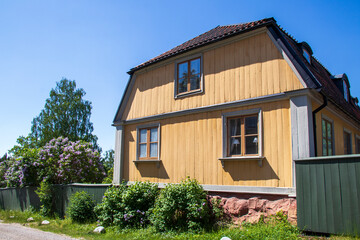 A yellow wooden house with a red tiled roof, green fence, and lush garden under a blue sky in Stockholm