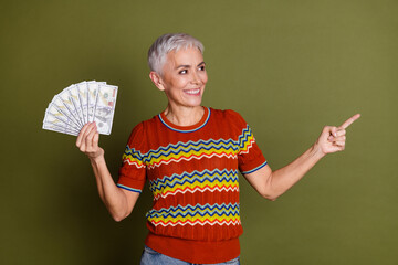 Charming senior woman holding money while pointing to the side against a khaki background,...
