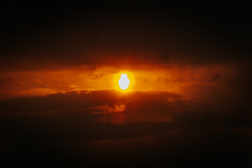 A partial solar eclipse glows through dramatic red-orange clouds, casting an intense and surreal light in a dark sky. Includes ample copy space.