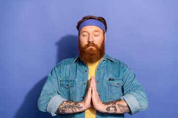 Relaxed and happy bearded man meditating in denim attire against a vibrant purple background with a peaceful expression and hands in prayer