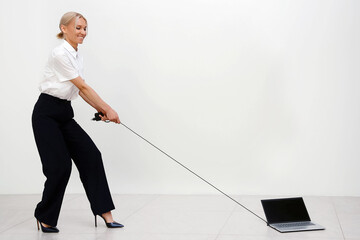 Smiling businesswoman in white shirt and black pants playfully pulls laptop by its power cord. She stands in clean, modern space, creating fun and unusual scene that mixes technology with humor