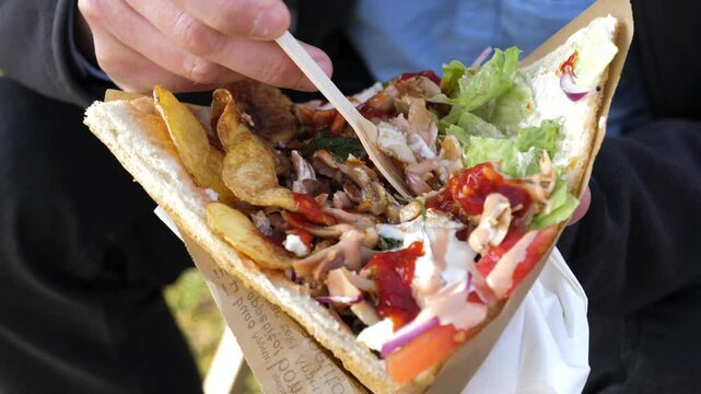 A man appetizingly holds and eats a kebab in a bread pita filled with chicken, vegetables and fried potato rings