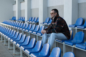A man sits alone on blue bleachers in a sports facility, gazing thoughtfully into the distance...