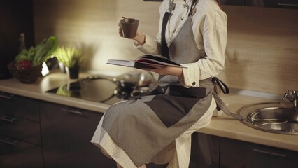 Woman chef reading cookbook and drinking coffee in kitchen. Female chef sitting on kitchen counter reading cookbook and drinking coffee, preparing for cooking