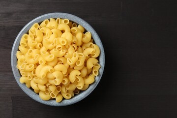 Raw horns pasta in bowl on dark wooden table, top view. Space for text