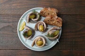 Delicious herring rolls with olives, pickles, dill and bread pieces on wooden table, top view