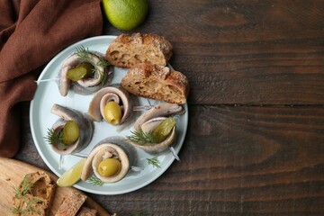 Delicious herring rolls with olives, pickles, dill and bread pieces on wooden table, flat lay....