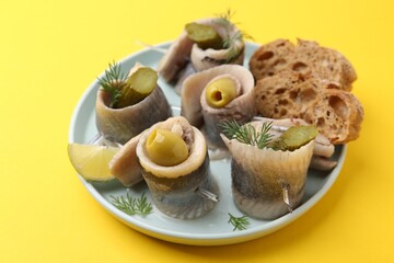 Delicious herring rolls with olives, pickles, dill and bread pieces on yellow table, closeup