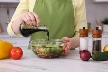 Woman pouring balsamic vinegar onto tasty salad at white marble table in kitchen, closeup