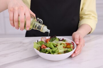 Woman pouring oil onto tasty salad at white marble table indoors, closeup