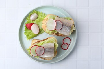 Tasty sandwiches with herring, radish and lettuce on white tiled table, top view
