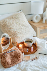 Cup of hot coffee on wicker rattan tray, boucle cosmetic bag, cosmetics on white bed linen.