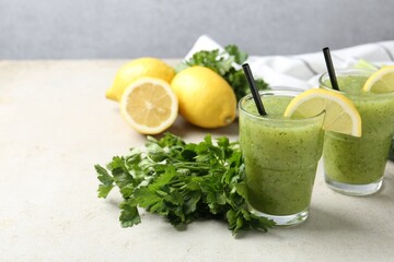 Healthy parsley smoothie in glasses, leaves and lemon on light textured table against grey background, closeup. Space for text