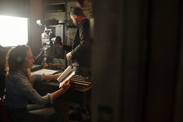 Young woman in headphones looking through text in paper documents while sitting by sound mixing...