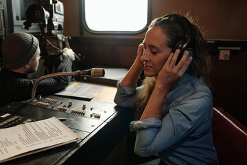 Young serene woman listening to pleasant music in headphones while sitting by sound mixing console...