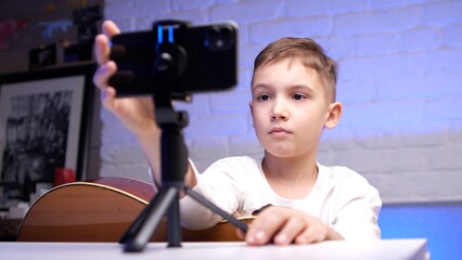 Young boy is recording himself playing guitar using a smartphone on a tripod. He is sitting at a...
