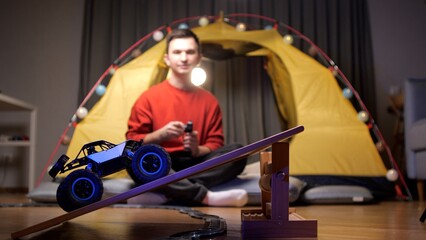 Man happily plays with a remote-controlled car in a cozy living room, driving it on a wooden ramp...