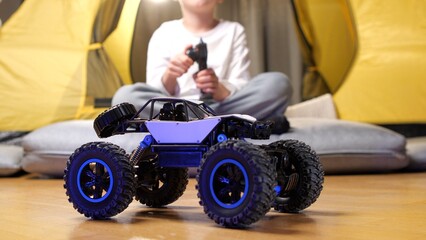Radio-controlled car standing on a wooden floor. A child is sitting in a tent, holding a remote control and playing with a toy car