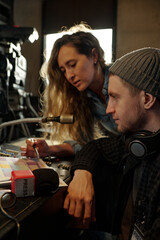 Young man sitting by console with microphone and helping female colleague with preparation for...