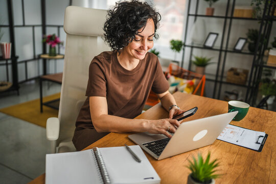 mature curly hair woman work from home on mobile phone and laptop