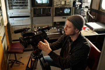 Young man adjusting focus of professional video camera while sitting by workplace with microphone...