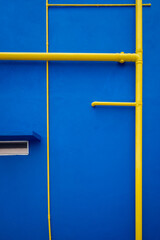 An abstract image of a bright blue wall with yellow pipes and conduits running across it, a small horizontal window on the left side